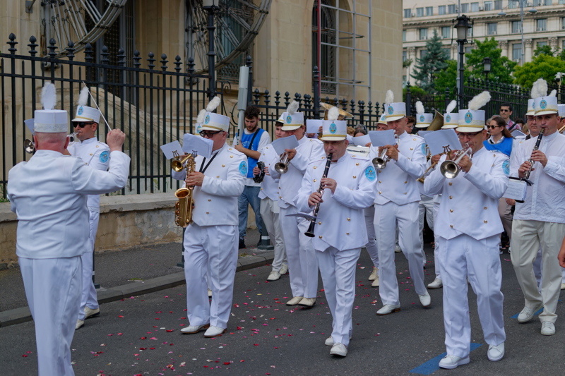 Procession with a marching brass band, featuring musicians wearing white uniforms and matching hats with white feathers